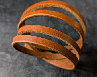 May include: A stacked leather bracelet in a warm brown hue. The bracelet features multiple thin, rounded leather bands, creating a layered effect. The leather has a natural grain and texture, suggesting a handcrafted item.