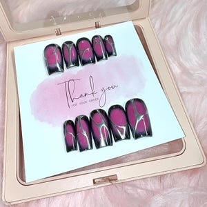 Puede incluir: Un conjunto de uñas postizas con un diseño negro, rosa y plateado. Las uñas se muestran en una tarjeta con las palabras "Thank you for your order" escritas en cursiva. Las uñas están en un estuche rosa.