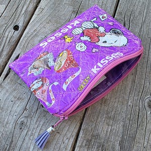 May include: A quilted purple zippered pouch featuring a Snoopy and Hershey's Kisses graphic print. The pouch has a pink zipper closure with a matching purple tassel pull. The exterior material has a shiny, crinkled texture with white stitching.