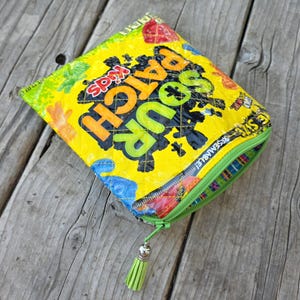 May include: A rectangular zippered pouch crafted from a repurposed yellow Sour Patch Kids candy bag. The exterior features the black and orange Sour Patch Kids logo, green and blue accents, and a quilted texture. A green tassel is attached to the zipper.