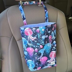 May include: A hanging car trash bag featuring a vibrant jellyfish print in shades of pink, blue, and purple against a black background. The bag has a top strap with snap closures and a light purple interior lining with a small rectangular label.