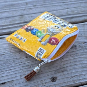 Peut inclure: Petite pochette zippée jaune fabriquée à partir d'emballages de bonbons recyclés. La pochette présente une fermeture éclair blanche, un pompon marron et un motif avec des bonbons colorés. Le texte "scan for something sweet" est visible.