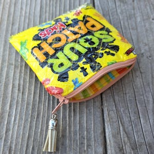 May include: A quilted zipper pouch crafted from a yellow Sour Patch Kids candy wrapper. The pouch features a pink zipper closure with a silver-toned metal tassel pull. The interior is lined with a multi-colored striped fabric in shades of green, yellow, and orange.