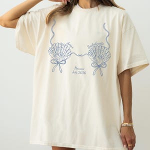Könnte beinhalten: Cremefarbenes T-Shirt mit blauem Muscheldesign. Das Design umfasst zwei Muscheln, die durch eine Perlenkette verbunden sind, mit dem Text "Amy's Bachelorette Hawaii July 2026."