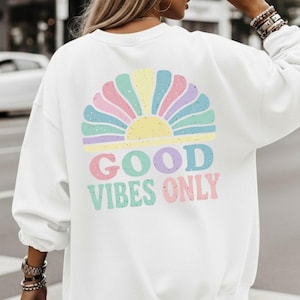 Könnte beinhalten: Weißes Sweatshirt mit einem farbenfrohen Sonnenaufgangsdesign auf dem Rücken. Der Sonnenaufgang hat Pastellfarben und die Worte "GOOD VIBES ONLY" in einer verspielten Schriftart. Ein lässiges und bequemes Oberteil.