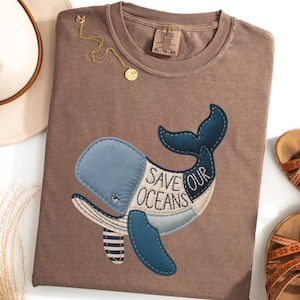 Könnte beinhalten: Ein braunes Comfort Colors T-Shirt mit einem Wal-Applikation. Der Wal ist blau und weiß mit den Worten "Save Our Oceans". Eine goldene Halskette und Sandalen sind ebenfalls sichtbar.