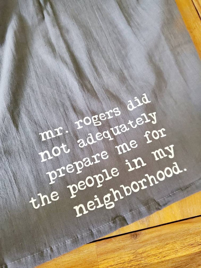 Pu&ograve; includere: Un asciugamano da cucina grigio con testo bianco che dice "mr. rogers did not adequately prepare me for the people in my neighborhood."