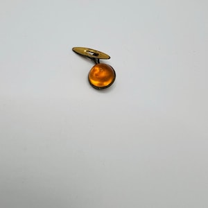 May include: A single vintage cufflink with a round, amber-colored stone set in a gold-toned metal frame. The cufflink has a small, flat, oval-shaped backing. The design is simple and elegant, suitable for formal wear.
