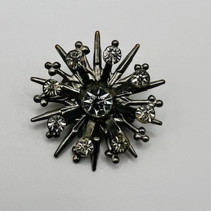 Puede incluir: Broche vintage plateado con diseño de estrella. El broche presenta un gran brillante central transparente rodeado de brillantes más pequeños y brillantes. El broche tiene un estilo clásico y elegante.