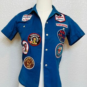 Op de afbeelding: Een blauw overhemd met korte mouwen en knoopsluiting, versierd met diverse geborduurde patches, waaronder 'BANTAM 100 GAME AJBC', 'PERFECT ATTENDANCE', 'TEAM CAPTAIN', 'AJBC MEMBER', 'I BEAT MY COACH' en 'I BEAT SANTA' met een afbeelding van de Kerstman.