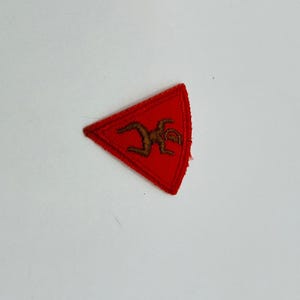 以下が含まれることがあります： 中央に茶色の刺繍が施された三角形の赤いワッペン。ワッペンには補強されたステッチの縁取りがあり、平らな布製で、手足を曲げた姿勢の様式化された人間のような形が描かれています。