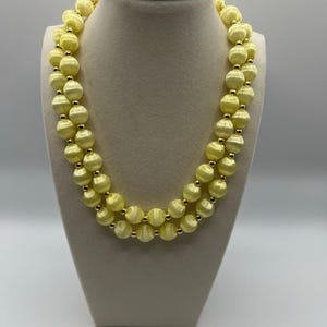 May include: A double-strand necklace featuring large, round, pale yellow beads. The beads are separated by small, gold-colored spacer beads. The necklace is displayed on a cream-colored bust.