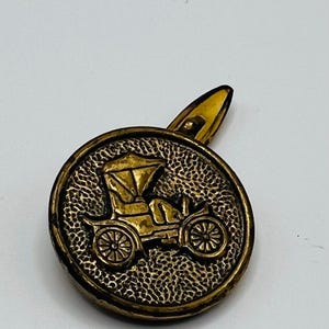 May include: Antique gold-colored tie clip with a detailed embossed design of an early automobile. The clip has a textured, circular face with a classic car image and a simple, pointed clasp.