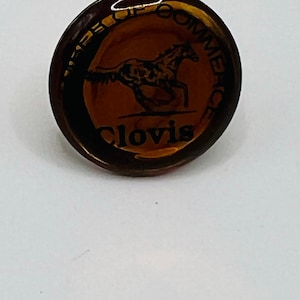 Puede incluir: Un pin redondo de color ámbar con el texto "Clovis" en la parte inferior. El pin presenta un gráfico de un caballo corriendo en el centro y las palabras "Chamber of Commerce" alrededor del borde.