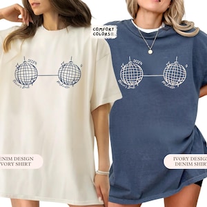 May include: Two t-shirts, one cream and one denim blue, each with a graphic of two disco balls linked by a line. The text "Olivia's Bach" and "Nashville" are below the disco balls. The cream shirt has "Denim Design Ivory Shirt" text.