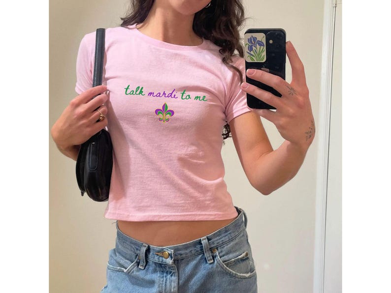 K&ouml;nnte beinhalten: Hellrosa T-Shirt mit dem gr&uuml;nen Schriftzug "talk mardi to me" und einer Fleur-de-Lis-Grafik in Lila, Gr&uuml;n und Gold. Das Shirt ist im Cropped-Stil gehalten und hat kurze &Auml;rmel.