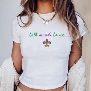 K&ouml;nnte beinhalten: Wei&szlig;es T-Shirt mit dem Schriftzug "talk mardi to me" in gr&uuml;ner Schrift &uuml;ber einem Fleur-de-Lis-Emblem in Lila, Gr&uuml;n und Gold. Das Shirt ist ein Cropped-Style, perfekt f&uuml;r den l&auml;ssigen Gebrauch.
