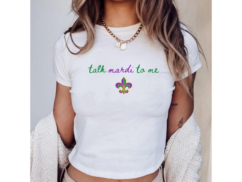 K&ouml;nnte beinhalten: Wei&szlig;es T-Shirt mit dem Schriftzug "talk mardi to me" in gr&uuml;ner Schrift &uuml;ber einem Fleur-de-Lis-Emblem in Lila, Gr&uuml;n und Gold. Das Shirt ist ein Cropped-Style, perfekt f&uuml;r den l&auml;ssigen Gebrauch.