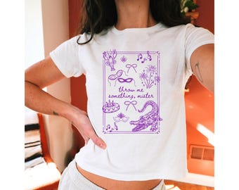 May include: White t-shirt with a purple graphic featuring a lobster, musical notes, masks, and an alligator. The text "throw me something, mister" is centered within a rectangular border.