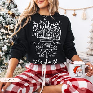 Pu&ograve; includere: Una felpa nera a maniche lunghe con una grafica bianca di un'auto che trasporta un albero di Natale, con il testo Our First Christmas, Est. 2026, Just Married e The Smith. Il design include un'auto con barattoli legati al paraurti posteriore.