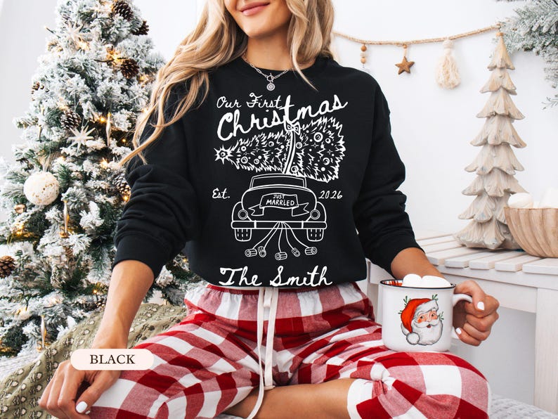 Pu&ograve; includere: Una felpa nera a maniche lunghe con una grafica bianca di un'auto che trasporta un albero di Natale, con il testo Our First Christmas, Est. 2026, Just Married e The Smith. Il design include un'auto con barattoli legati al paraurti posteriore.