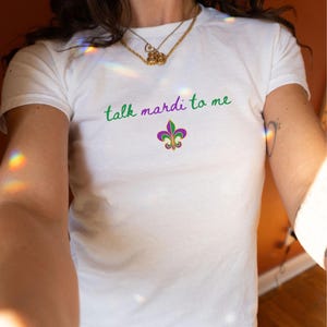 K&ouml;nnte beinhalten: Wei&szlig;es T-Shirt mit dem Schriftzug "talk mardi to me" in gr&uuml;ner und violetter Schrift. Unter dem Text befindet sich eine Fleur-de-Lis-Grafik in Lila, Gr&uuml;n und Gold. Das T-Shirt hat einen Rundhalsausschnitt.