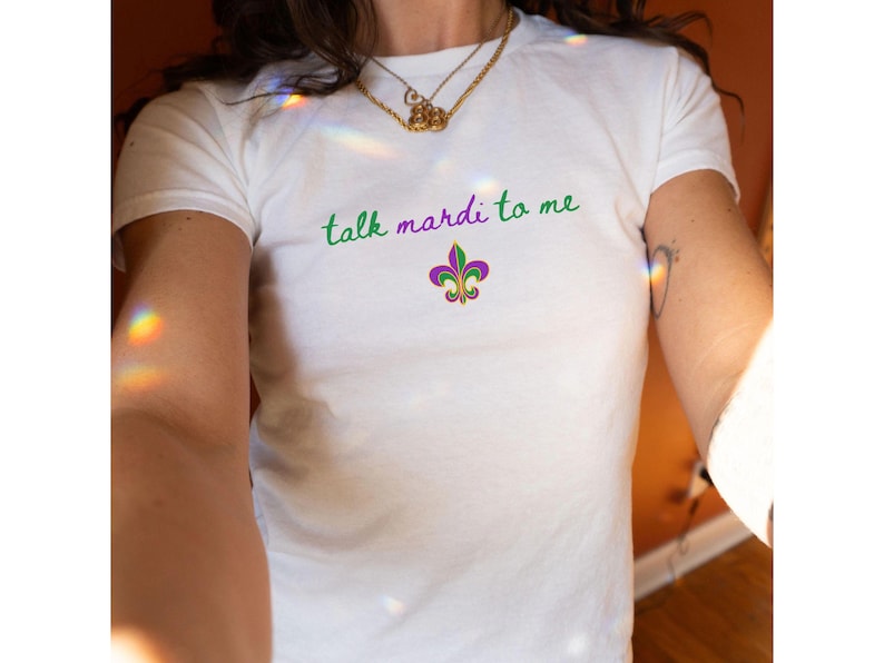 K&ouml;nnte beinhalten: Wei&szlig;es T-Shirt mit dem Schriftzug "talk mardi to me" in gr&uuml;ner und violetter Schrift. Unter dem Text befindet sich eine Fleur-de-Lis-Grafik in Lila, Gr&uuml;n und Gold. Das T-Shirt hat einen Rundhalsausschnitt.