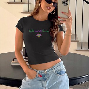 K&ouml;nnte beinhalten: Schwarzes T-Shirt mit Rundhalsausschnitt und dem Text "talk mardi to me" in Gr&uuml;n sowie einer Fleur-de-Lis-Grafik. Das Shirt hat eine figurbetonte Passform mit kurzen &Auml;rmeln. Das Model tr&auml;gt eine hellblaue Jeans.