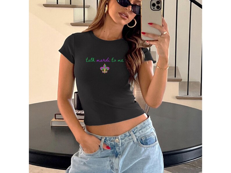 K&ouml;nnte beinhalten: Schwarzes T-Shirt mit Rundhalsausschnitt und dem Text "talk mardi to me" in Gr&uuml;n sowie einer Fleur-de-Lis-Grafik. Das Shirt hat eine figurbetonte Passform mit kurzen &Auml;rmeln. Das Model tr&auml;gt eine hellblaue Jeans.