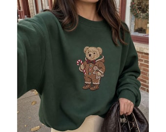 May include: A dark forest green crewneck sweatshirt featuring a central embroidered graphic of a teddy bear wearing a brown gingerbread-style hooded onesie, a red and green bowtie, and holding a red and white striped candy cane and a cookie.
