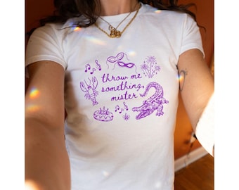May include: White t-shirt with a purple graphic design. The design includes a crawfish, musical notes, a mask, fireworks, a cake, a fleur-de-lis, and an alligator. The text "throw me something, mister" is also included.