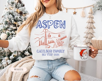 May include: White sweatshirt with a blue "ASPEN" graphic and a red ski lift and mountain design. Below the graphic is "CARLSON FAMILY SKI TRIP 2026". A white mug with a Santa Claus graphic is held in the hand.