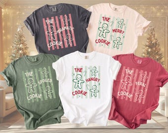 May include: Five short-sleeve t-shirts in charcoal, cream, olive green, white, and red. Each shirt features a central graphic with vertical stripes, gingerbread men, candy canes, and text including The Dramatic Cookie, The Merry Cookie, The Spoiled Cookie, The Hangry Cookie, and The Feral Cookie.