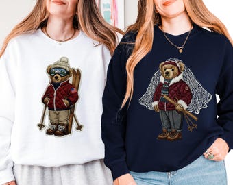 May include: Two sweatshirts, one white and one navy blue, each featuring a detailed graphic of a bear in winter attire. The bear wears a quilted jacket, hat, and ski goggles, holding skis. The white sweatshirt has the bear with an American flag.