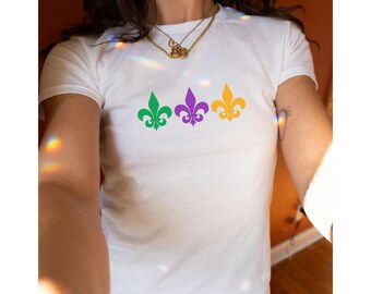 May include: White t-shirt featuring three fleur-de-lis symbols in green, purple, and gold. The symbols are arranged horizontally across the chest. The shirt is made of a soft material and is suitable for casual wear.