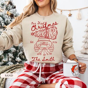 Pu&ograve; includere: Una felpa girocollo color sabbia con una stampa grafica rossa. Il design mostra un'auto che trasporta un albero di Natale con il testo "Our First Christmas", "Est. 2026", "Just Married" e "The Smith" stampato in carattere corsivo.