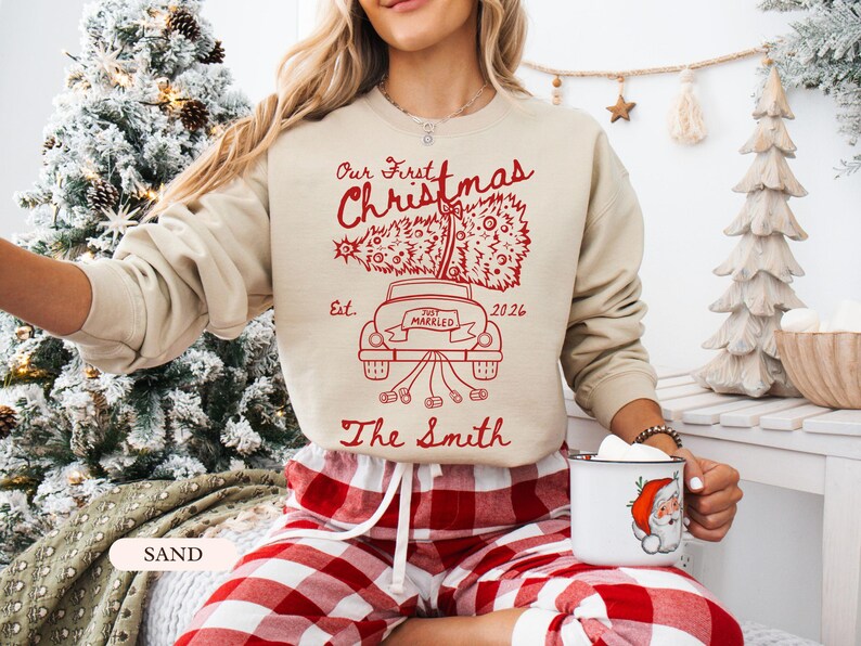 Pu&ograve; includere: Una felpa girocollo color sabbia con una stampa grafica rossa. Il design mostra un'auto che trasporta un albero di Natale con il testo "Our First Christmas", "Est. 2026", "Just Married" e "The Smith" stampato in carattere corsivo.