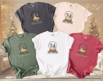 May include: Five short-sleeve crew neck t-shirts in charcoal, light pink, olive green, white, and maroon. Each shirt features a graphic of a snow globe containing a snowman and a Christmas tree, with the text "THE SMITHS Holiday Cheer Department".