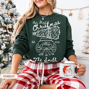 Pu&ograve; includere: Una felpa a maniche lunghe verde scuro con una grafica bianca di un'auto che trasporta un albero di Natale sul tetto. Il testo recita Our First Christmas, Est. 2026, Just Married e The Smith, con un fiocco e dei barattoli che pendono dietro.