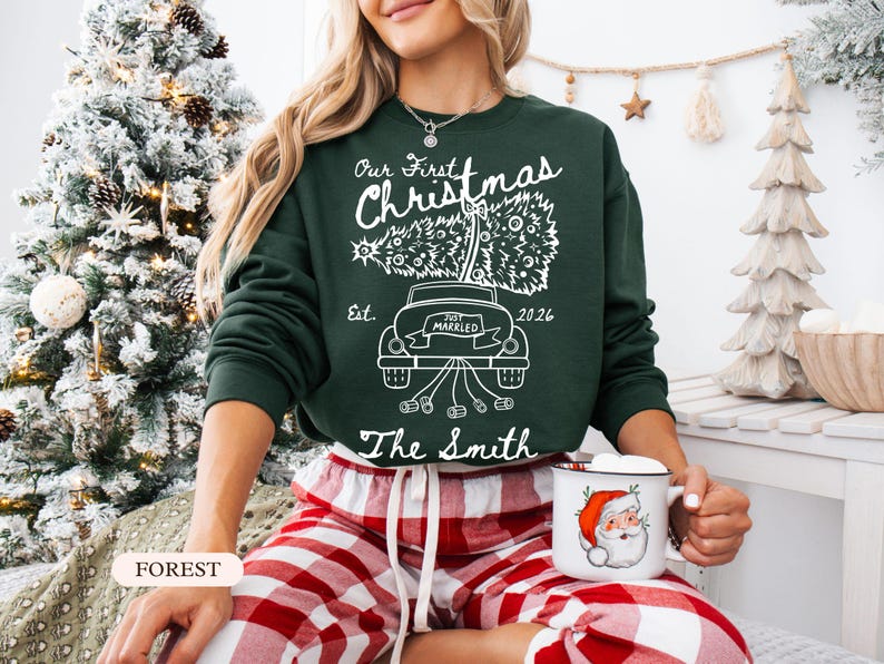 Pu&ograve; includere: Una felpa a maniche lunghe verde scuro con una grafica bianca di un'auto che trasporta un albero di Natale sul tetto. Il testo recita Our First Christmas, Est. 2026, Just Married e The Smith, con un fiocco e dei barattoli che pendono dietro.