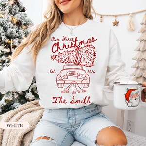 Pu&ograve; includere: Una felpa bianca a maniche lunghe con una stampa grafica rossa di un'auto che trasporta un albero di Natale sul tetto. Il testo recita Our First Christmas, Est. 2026, Just Married e The Smith, con lattine legate al paraurti posteriore.