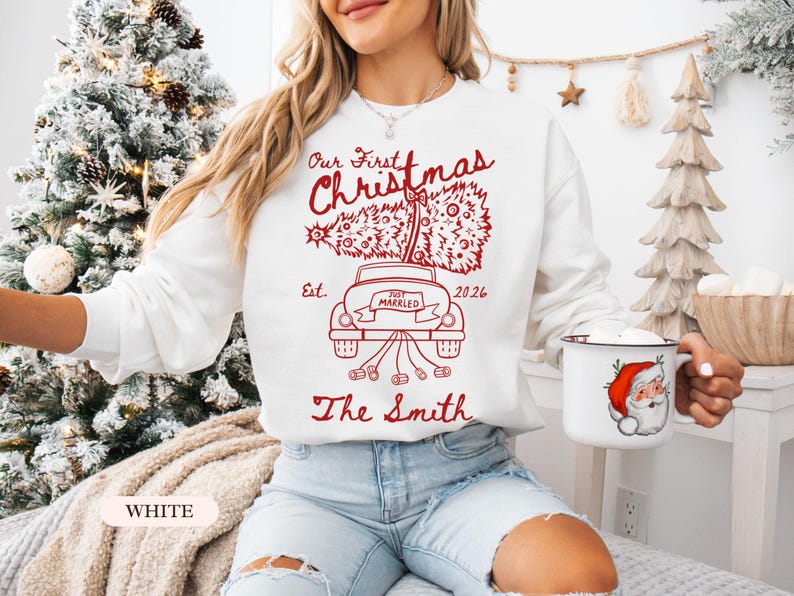Pu&ograve; includere: Una felpa bianca a maniche lunghe con una stampa grafica rossa di un'auto che trasporta un albero di Natale sul tetto. Il testo recita Our First Christmas, Est. 2026, Just Married e The Smith, con lattine legate al paraurti posteriore.