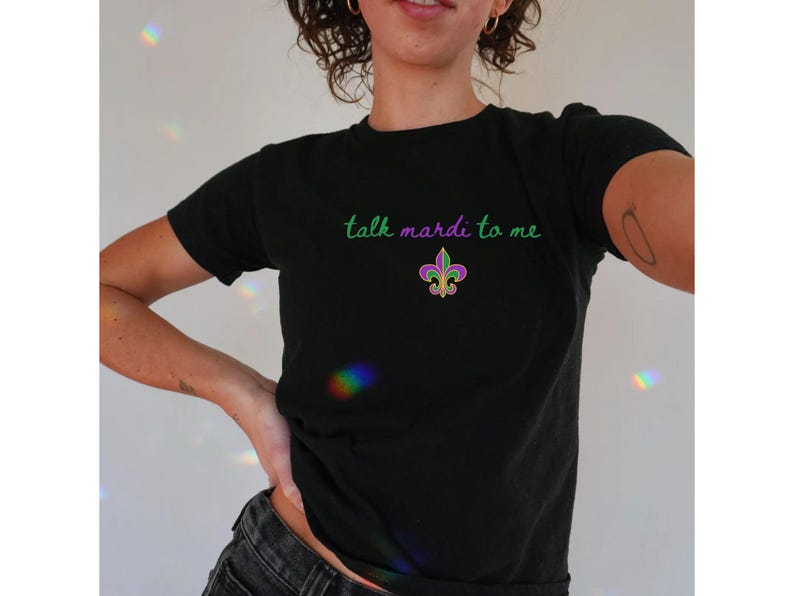 K&ouml;nnte beinhalten: Schwarzes T-Shirt mit dem Schriftzug "talk mardi to me" in gr&uuml;ner Schrift und einem Fleur-de-Lis-Design in Lila, Gr&uuml;n und Gold. Das T-Shirt hat einen Rundhalsausschnitt und scheint aus Baumwolle zu sein.