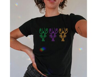 May include: Black t-shirt featuring three lobster illustrations in green, purple, and yellow. The lobsters are arranged horizontally across the front of the shirt. The shirt is a short-sleeved crew neck.