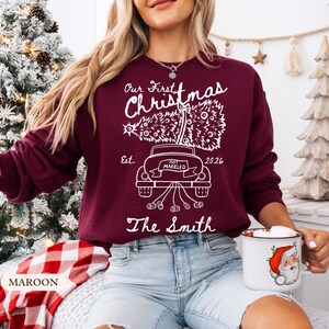 Pu&ograve; includere: Una felpa a maniche lunghe color bordeaux con una grafica bianca di un'auto che trasporta un albero di Natale. Il testo recita Our First Christmas, Est. 2026, Just Married e The Smith. Il design include un'auto con barattoli al seguito.