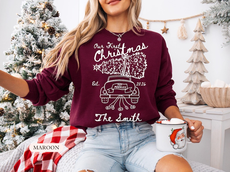 Pu&ograve; includere: Una felpa a maniche lunghe color bordeaux con una grafica bianca di un'auto che trasporta un albero di Natale. Il testo recita Our First Christmas, Est. 2026, Just Married e The Smith. Il design include un'auto con barattoli al seguito.
