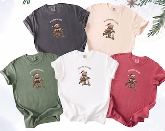 May include: Five short-sleeved crewneck t-shirts in charcoal, cream, sage green, white, and red. Each shirt features a graphic of a teddy bear wearing a Santa hat and a patterned sweater, riding a scooter, with unique holiday-themed text above.