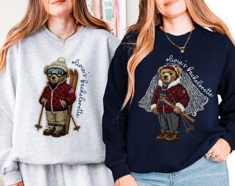 May include: Two sweatshirts, one light gray and one navy blue, each featuring a bear wearing ski gear. The bear on the gray sweatshirt has goggles and skis, while the bear on the navy sweatshirt wears a veil. Text reads "Olivia's Bachelorette".