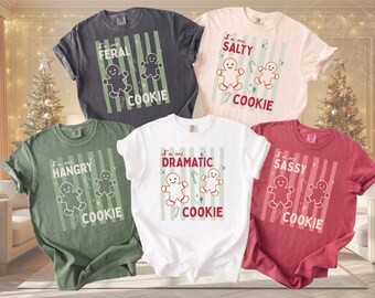 May include: Five short-sleeve t-shirts in charcoal, cream, olive green, white, and red. Each shirt features a graphic of two gingerbread people, vertical stripes, and text reading I'm one FERAL, SALTY, HANGRY, DRAMATIC, or SASSY COOKIE.