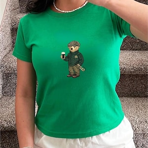Può includere: T-shirt verde con un orso ricamato che indossa un berretto di tweed, un maglione verde con un trifoglio e tiene in mano una pinta di stout. L'orso indossa anche pantaloni e scarpe marroni e tiene un paio di occhiali. La maglietta è girocollo.